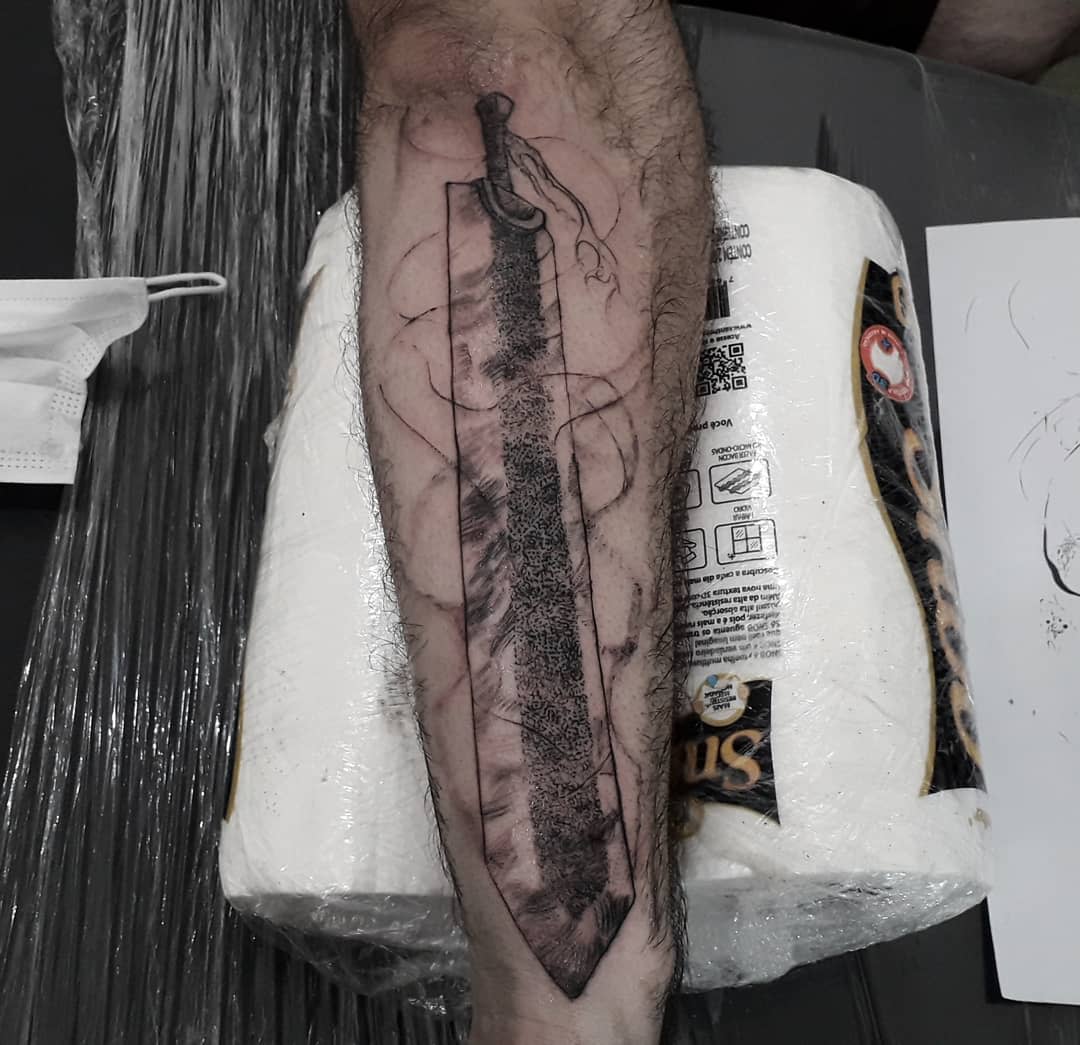 Black and gray tattoo of Guts' Dragon Slayer sword from Berserk on the forearm, depicted with a worn, battle-scarred texture and a flowing ribbon